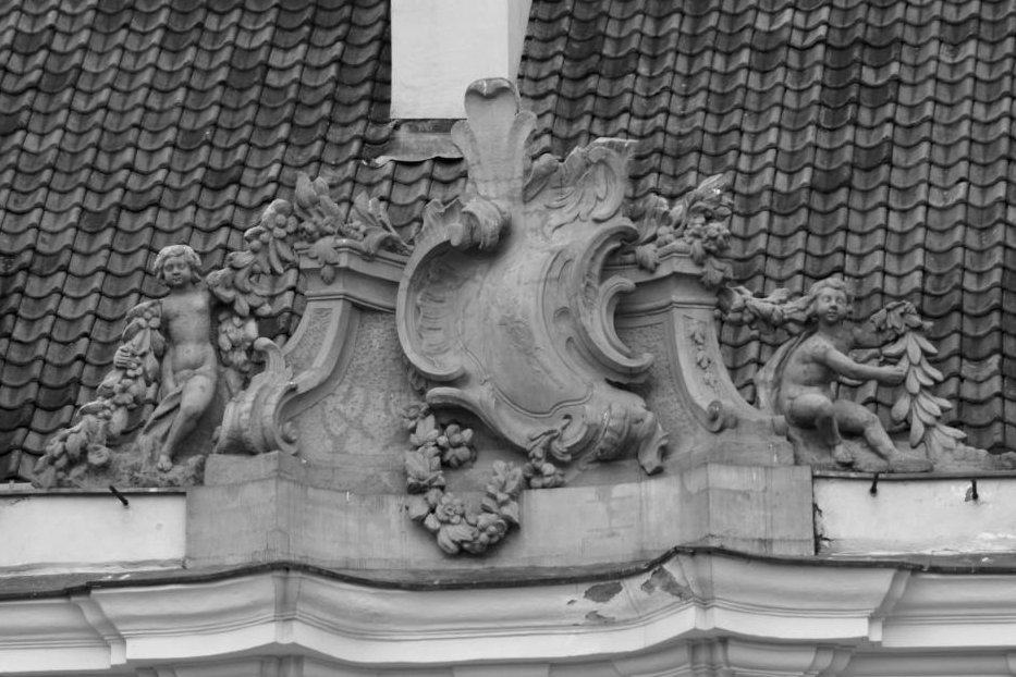 Czarno białe zdjęcie. Przedstawia fragment dekoracji kamienicy Prażmowskich w Warszawie. Jest to kartusz, z boku dwie postacie chłopców wśród kwiatów. 



