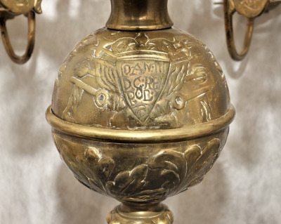 Zbliżenie na fragment żyrandola. Od dołu ozdobę stanowi kula dekorowana trzema emblematami: herbem Janina należącym do króla Jana III, tarczą z wizerunkiem orła oraz inskrypcją. 