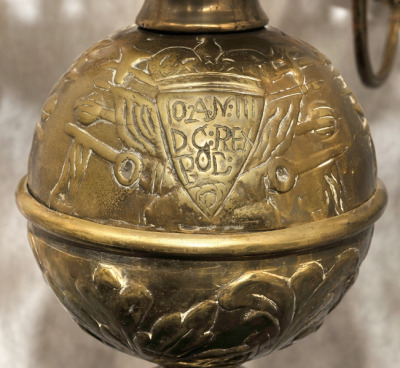 Zbliżenie na fragment żyrandola. Od dołu ozdobę stanowi kula dekorowana trzema emblematami: herbem Janina należącym do króla Jana III, tarczą z wizerunkiem orła oraz inskrypcją. 