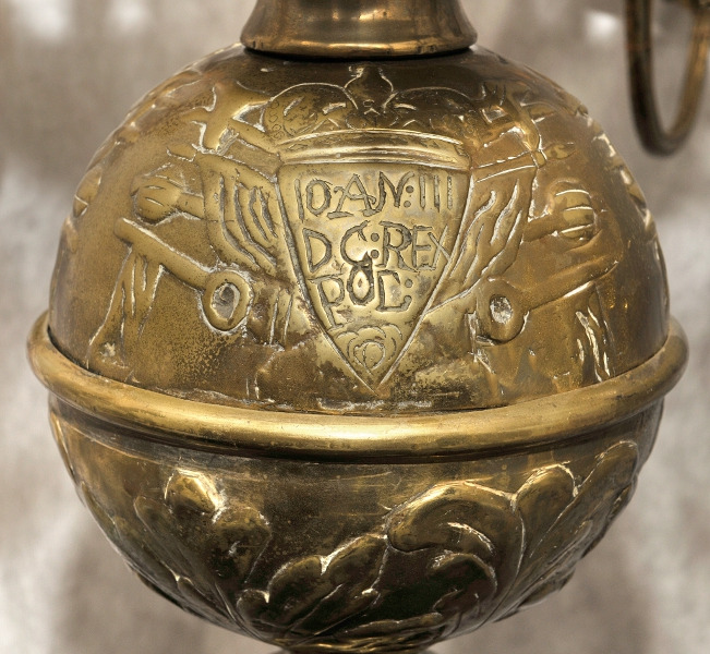 Zbliżenie na fragment żyrandola. Od dołu ozdobę stanowi kula dekorowana trzema emblematami: herbem Janina należącym do króla Jana III, tarczą z wizerunkiem orła oraz inskrypcją. 