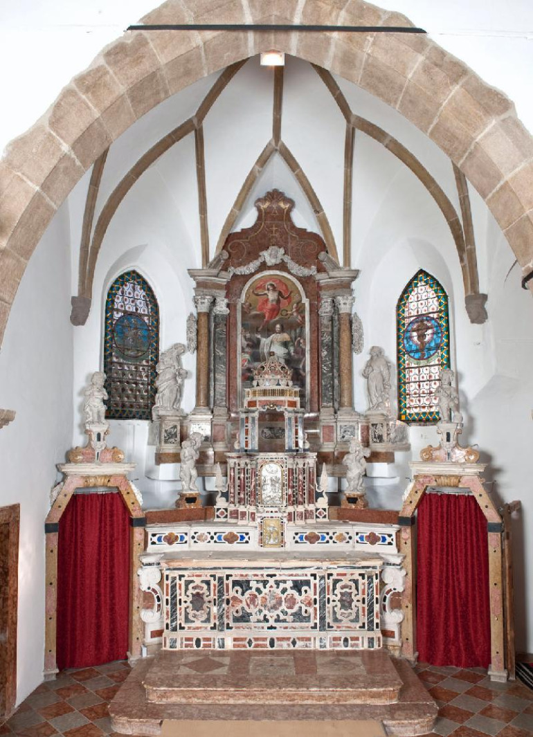 Zdjęcie przedstawia widok na cały ołtarz. Pośrodku tabernakulum i drzwi tabernakulum z wizerunkiem papieża, cesarza, Jana III i weneckiego doży. Po bokach rzeźby świętych. Pomiędzy dwoma oknami z witrażami obraz w barokowej oprawie architektonicznej.