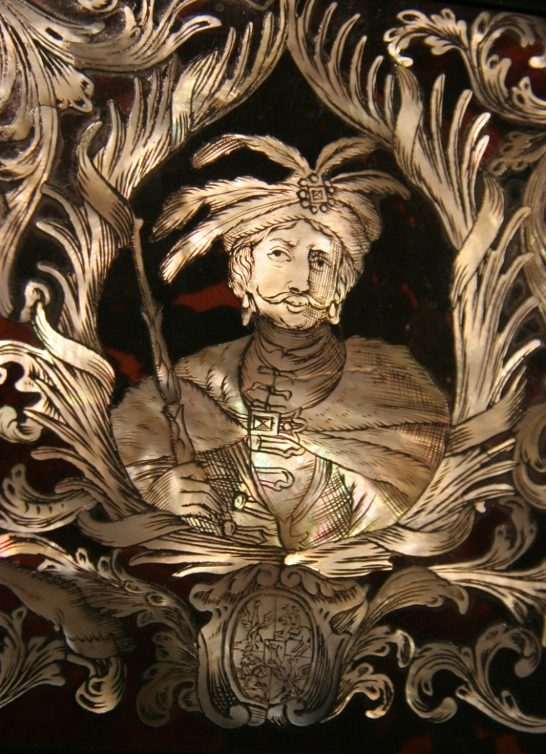 Zbliżenie na portret Jana III Sobieskiego ze stołu do gry Maksymiliana Emanuela. Król ukazany w stroju historycznym - żupanie i delii. Portret zamknięty w roślinne motywy. Pod wizerunkiem Jana III Sobieskiego herb.
