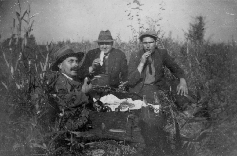 Czarno-biała fotografia przedstawia trzech mężczyzn, którzy siedzą na łonie natury, w trawie. Patrzą prosto w obiektyw. Odpoczywają i spożywają posiłek.