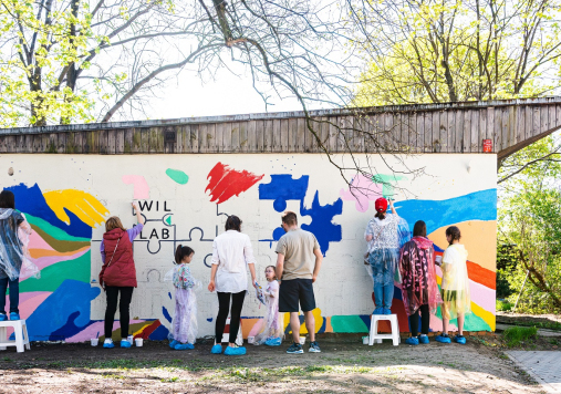 Ludzie malujący kolorowy mural na białej ścianie, napis: Wil.Lab 